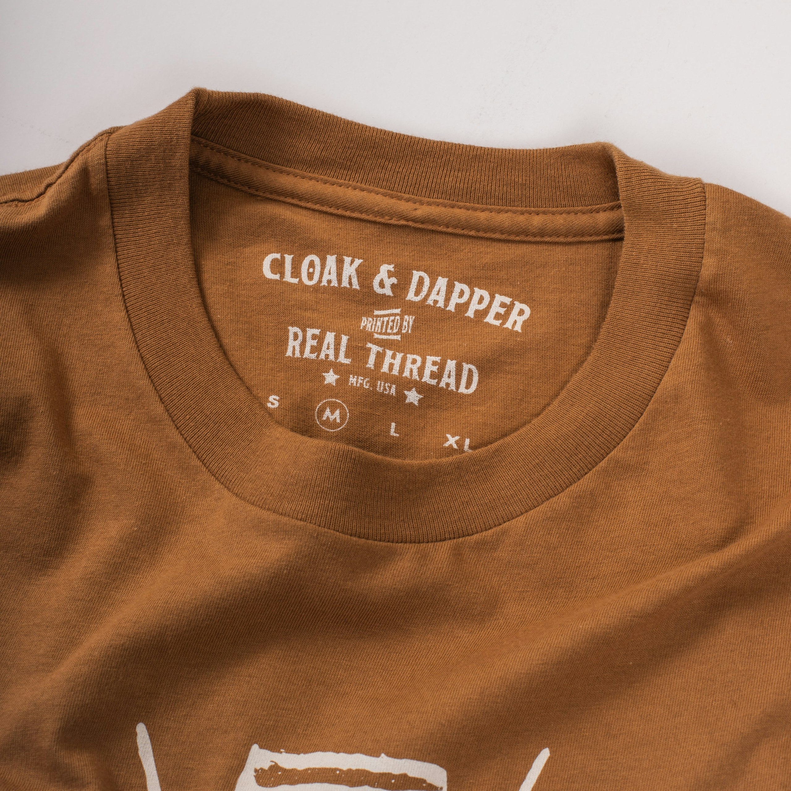 A burnt orange shirt with a custom printed tag that says "cloak and dapper, printed by Real Thread" 